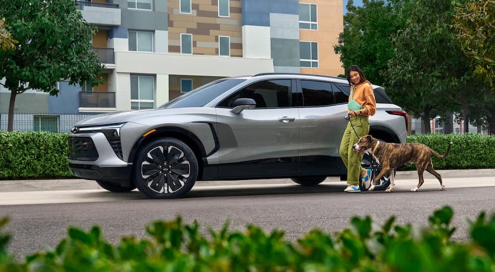 A grey 2025 Chevy Blazer EV RS is parked on the side of a city street as a woman and a dog jog by.