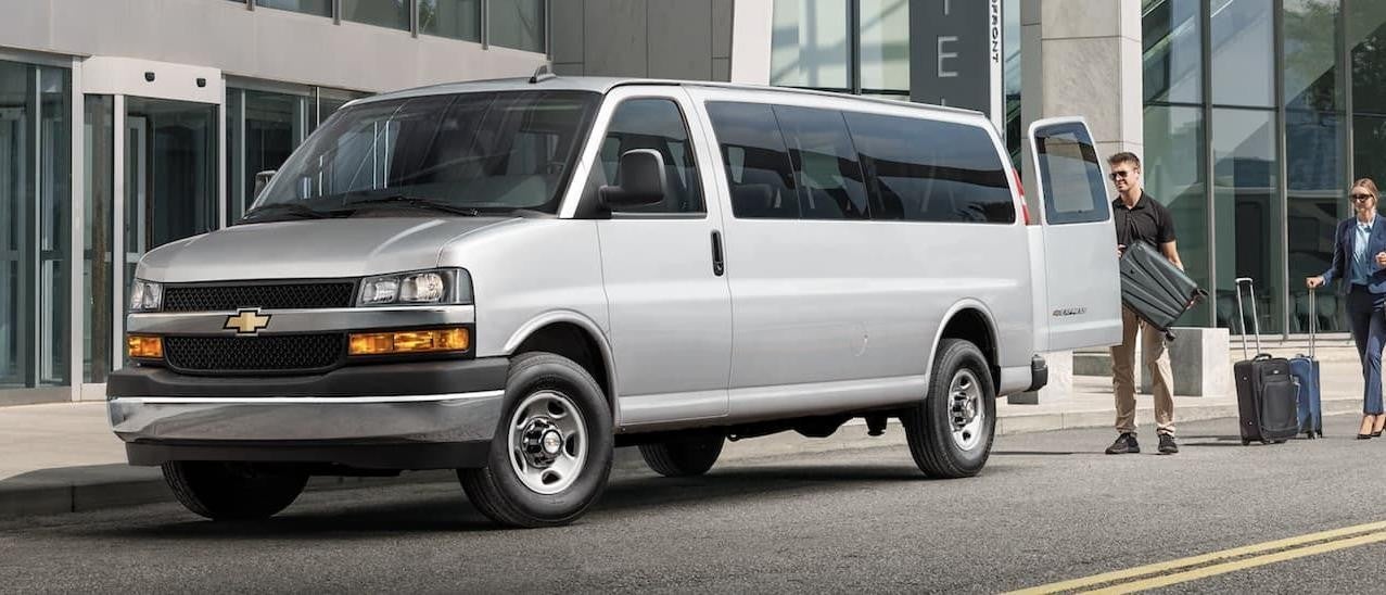 A silver 2022 Chevy Express Van is shown from the side as two people load suitcases into the vehicle.