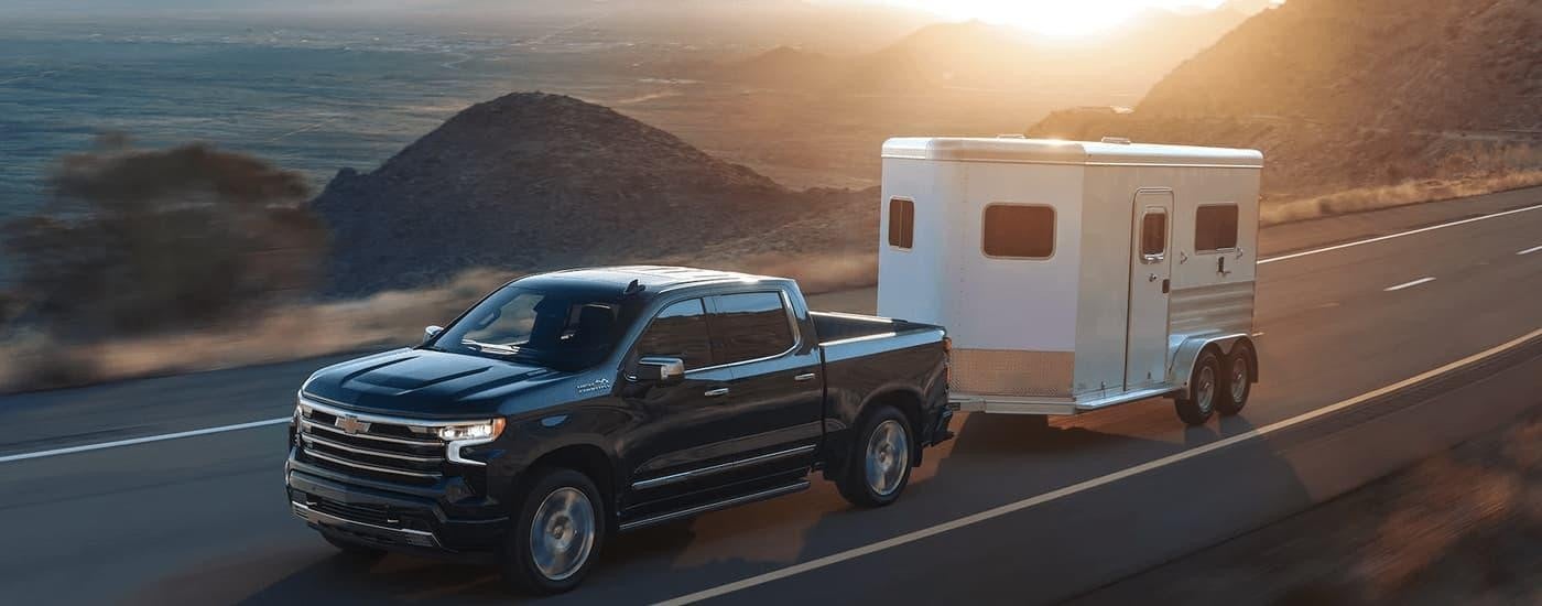 A grey 2023 Chevy Silverado 1500 High Country is shown towing a white trailer on an open road.
