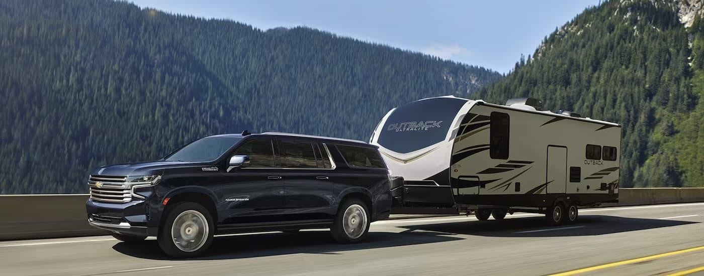A dark blue 2024 Chevy Suburban High Country is shown towing a camper trailer on a mountain road.