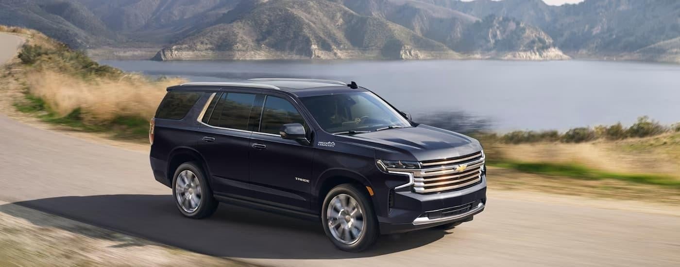 A blue 2024 Chevy Tahoe High Country is shown driving on an open road near a lake.
