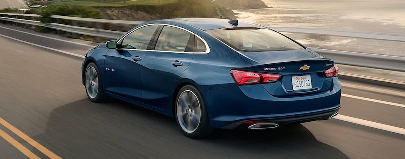 A blue 2023 Chevy Malibu is shown from a rear angle driving on a highway next to an ocean.