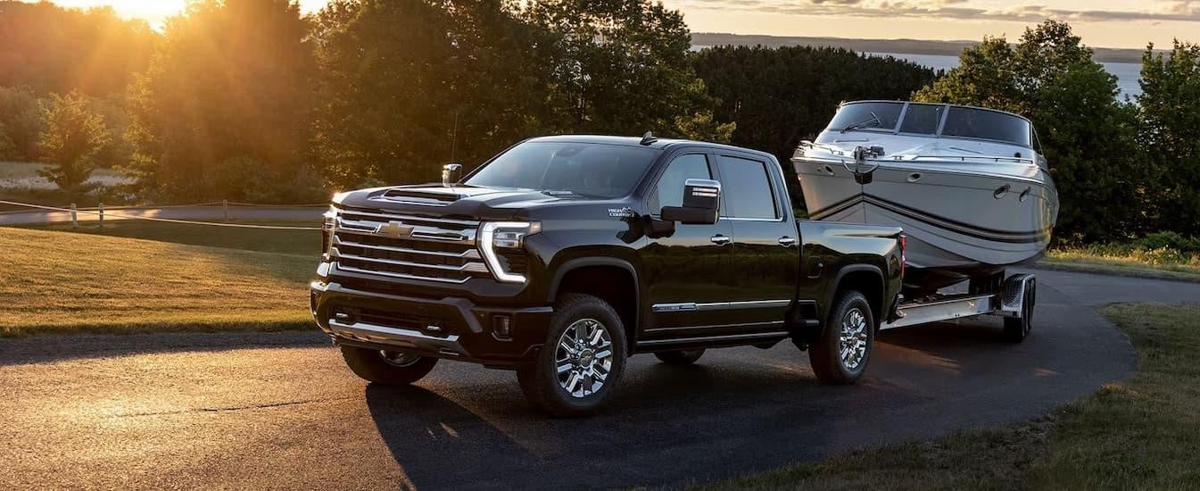 A black 2024 Chevy Silverado 2500HD High Country is shown towing a white boat on a winding road.