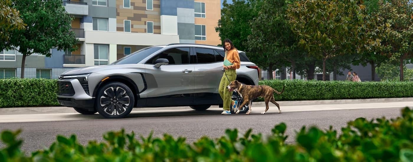 A silver 2025 Chevy Blazer EV RS is parked on the side of a city street as a woman walks by with her dog.